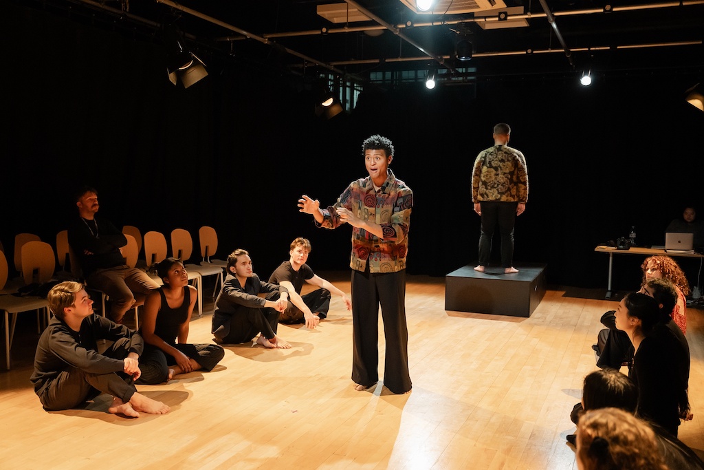 An actor stands between two groups of students seated on the stage, in mid-performance, while another actor stands behind him on a black riser with his back to the actor.